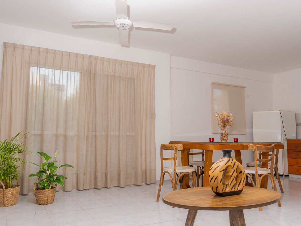 A dining area is presented with a wooden table and four chairs, accompanied by a natural wooden centerpiece. Light-colored curtains frame a large window, allowing natural light to fill the space. Potted plants are located near the window, adding a touch of greenery.