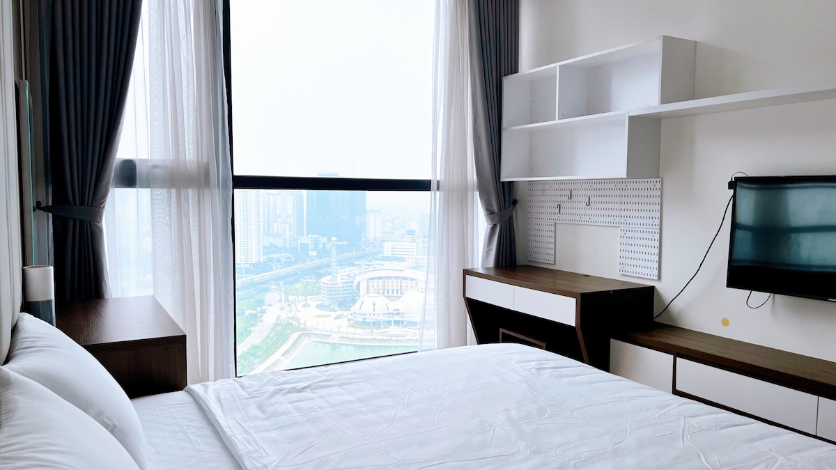 A bright bedroom features a spacious bed with crisp white linens, complemented by a wooden bedside table. Large windows offer expansive views of the city skyline. A wall-mounted television and a minimalist desk add functionality, while soft curtains filter natural light.