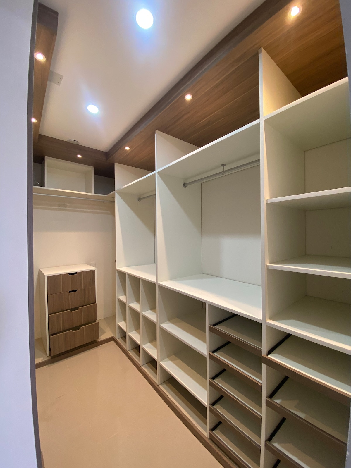 A spacious walk-in closet features multiple shelves and hanging rods. Furniture pieces including a tall dresser and various storage options are offered for organization. Soft overhead lighting illuminates the area, highlighting the clean lines and open layout.