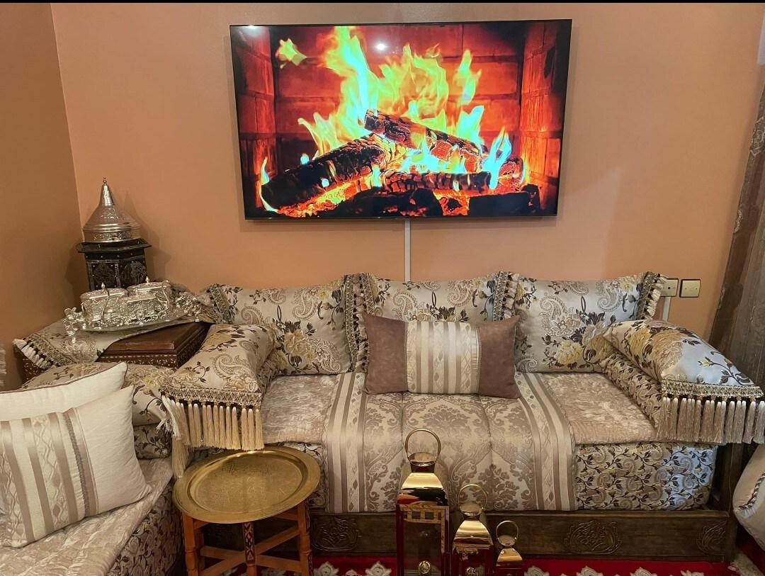 A plush sofa with intricate fabric patterns is positioned in front of a large television displaying a flickering fire. Decorative pillows and a small table with metallic accents complement the cozy seating area, offering a warm and inviting ambiance.