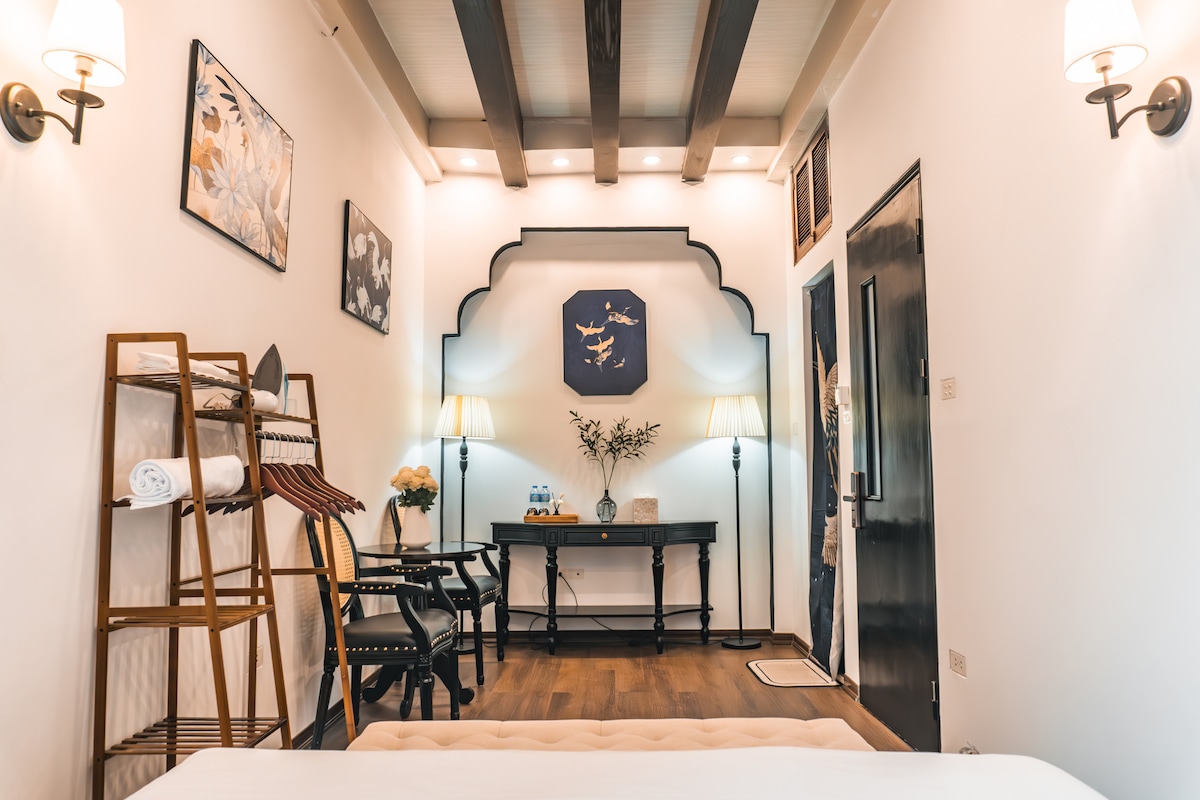 A well-appointed room features wooden beams overhead, with neutral walls and artistic décor. A small table with two chairs sits beside a floral arrangement. A towel rack with neatly folded towels is positioned against the wall near the door, allowing for easy access.
