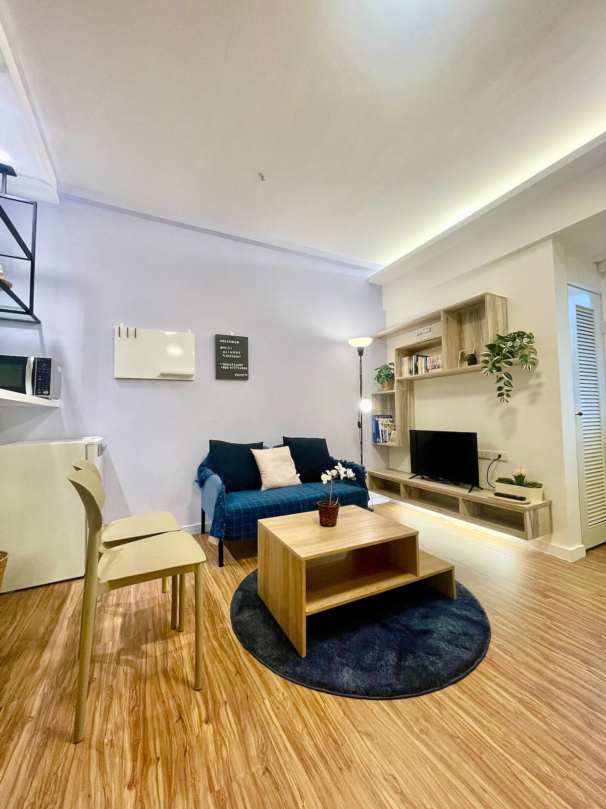A living area is presented with a cozy blue armchair adorned with cushions. A small coffee table is centered on a round rug, and a television is positioned on a wooden shelf. A plant adds a touch of greenery, complemented by soft overhead lighting.