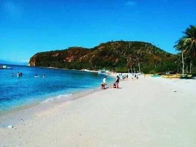 A sandy beach stretches along the coastline, with gentle waves lapping at the shore. Clear blue skies are overhead, and a lush green hill rises in the background. Various individuals are seen walking, swimming, and enjoying the serene environment.