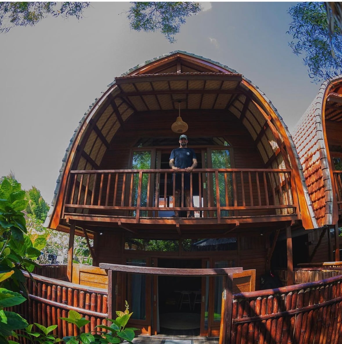 The exterior of a wooden bungalow is displayed, showcasing a unique arched roof design. A balcony extends from the second floor, framed by lush greenery. The entrance features wooden doors, and an inviting outdoor space is highlighted below.