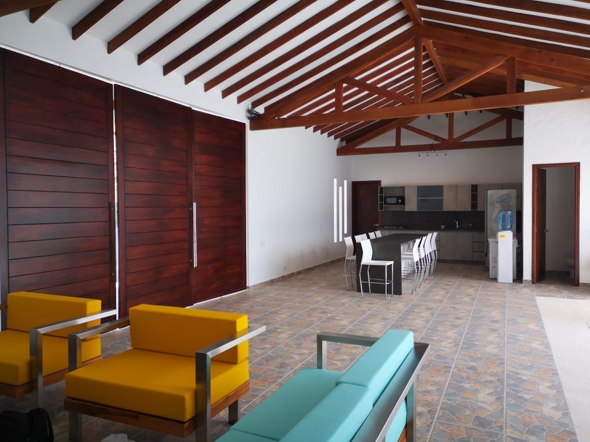 An open-plan living space features high wooden ceilings and large sliding doors, allowing natural light to fill the area. Modern seating in bright colors is arranged around a spacious dining table. The kitchen area is well-equipped, seen in the background.