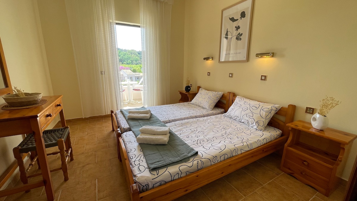 A well-lit room features a twin bed arrangement with patterned bedding and neatly folded towels. Natural light enters through a sliding door leading to a balcony. Two wooden nightstands stand beside the beds, and a small wooden desk complements the space.