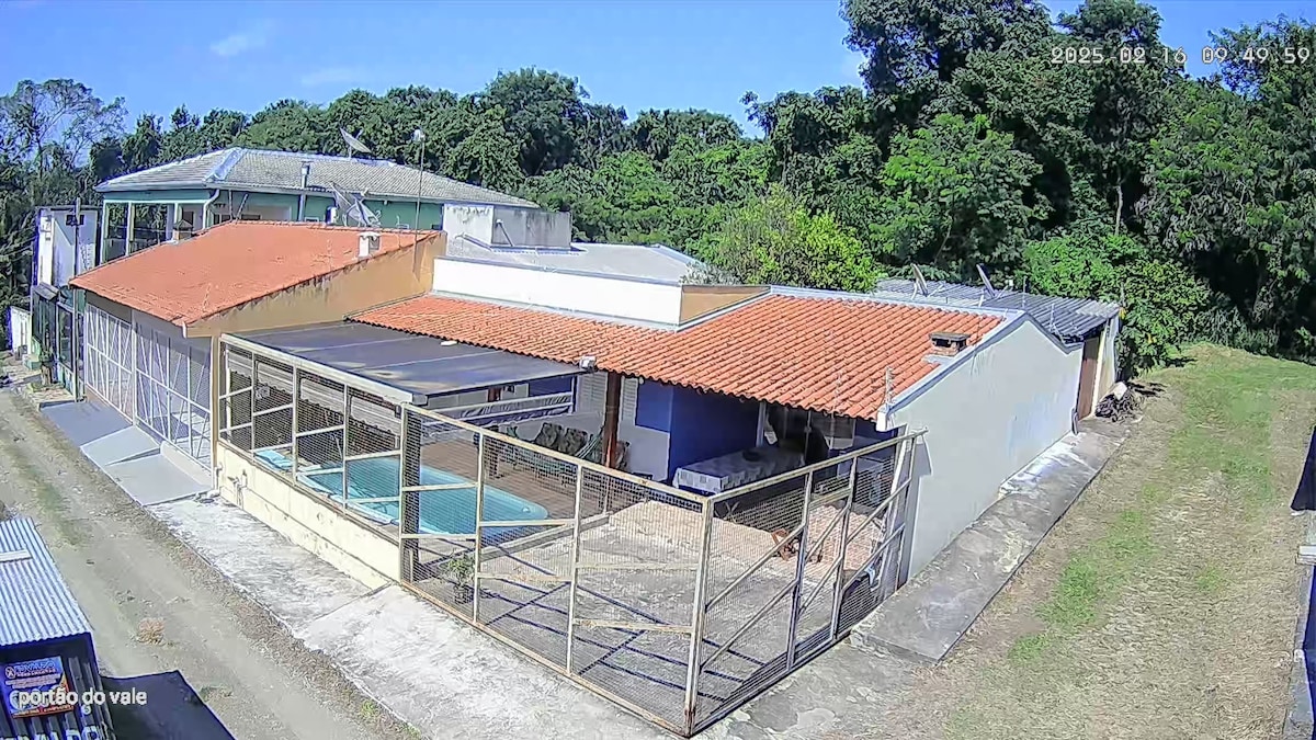 A spacious outdoor area is visible, featuring a fenced pool and a patio space. The surrounding greenery adds to the natural setting. The house displays a combination of flat and sloped roofs, with various windows allowing for ample daylight.