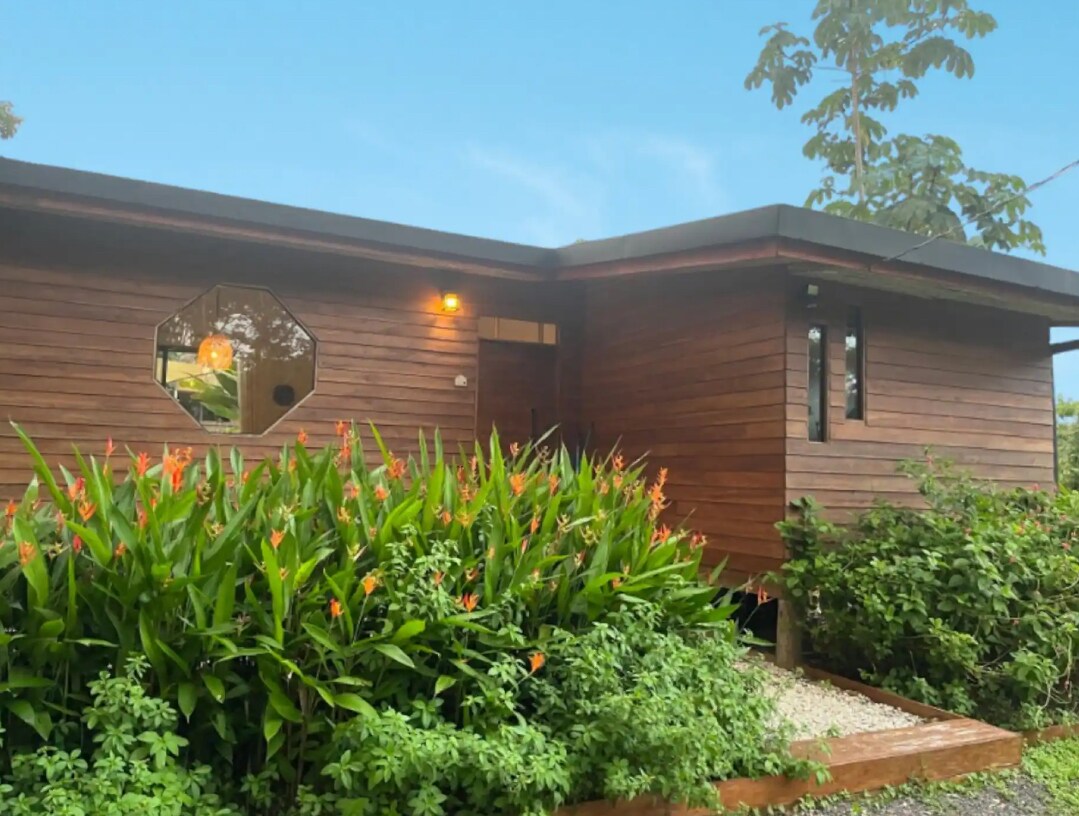 The exterior of a modern wooden villa is surrounded by lush tropical plants and vibrant flowers. An octagonal window adds architectural interest to the facade, while soft lighting provides a welcoming glow as dusk approaches. Gravel pathways lead up to the entrance, blending into the natural scenery.