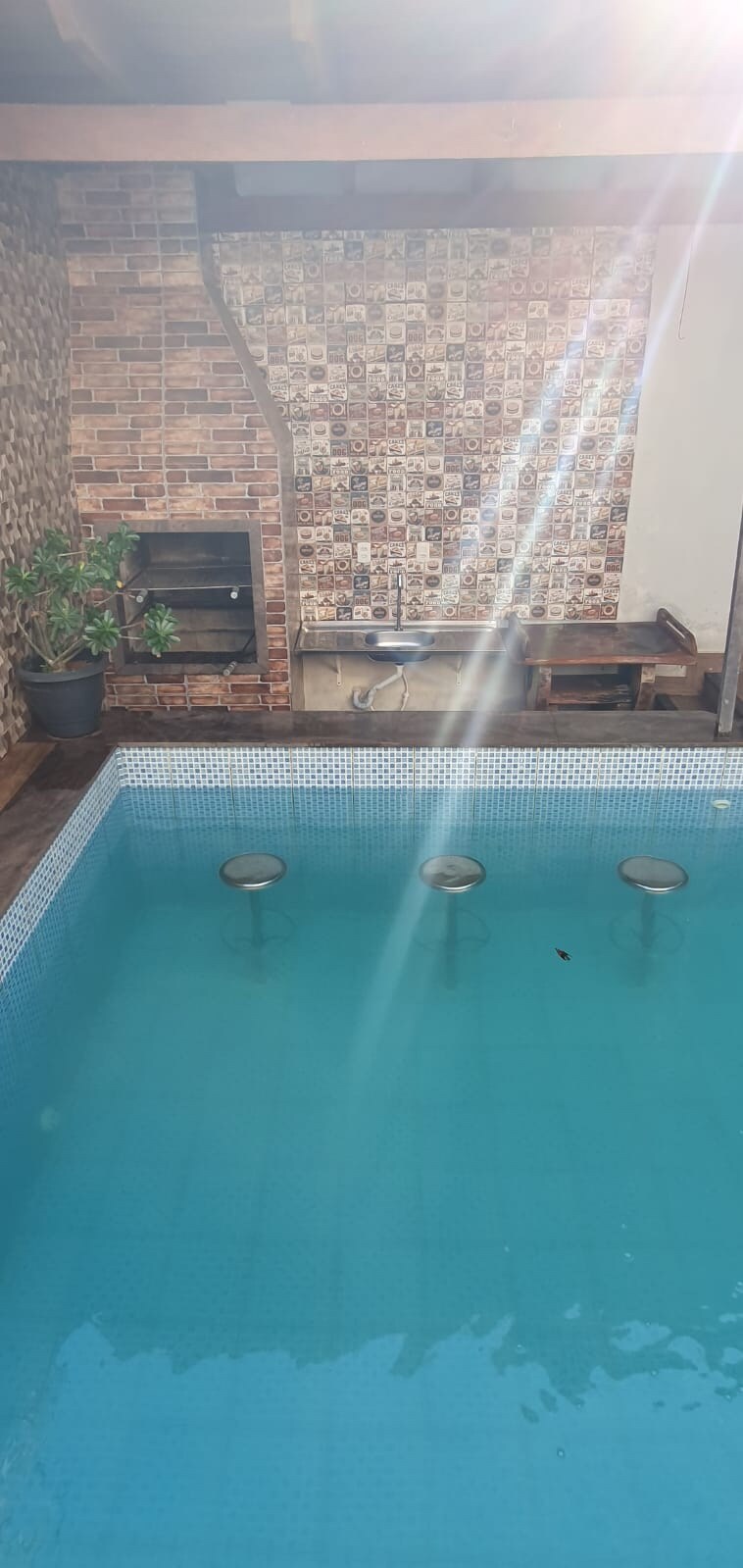 A swimming pool with clear blue water is featured, accompanied by sunken seating along the edge. A small barbecue area is visible in the background, accented by a textured brick wall. Natural light reflects off the water's surface, creating a serene environment.