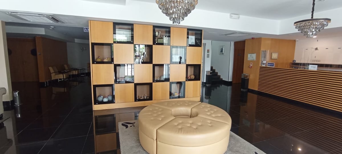 A spacious lobby area is highlighted by a circular, tufted seating arrangement in a neutral tone. A wooden shelving unit contains decorative items, while polished black tiles reflect the soft lighting from an elegant chandelier overhead.