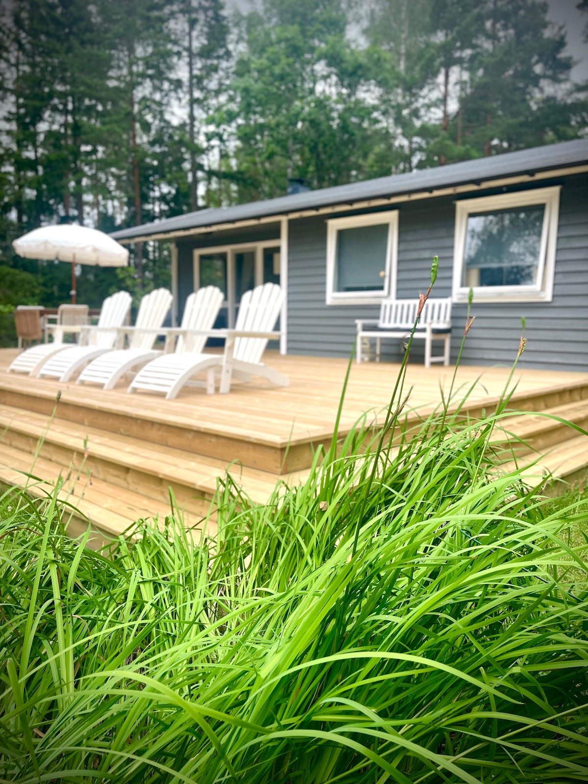 A wooden terrace is presented in the foreground, adorned with several lounge chairs and a white umbrella. The exterior of the house features grey siding, and large windows face a tranquil natural landscape of greenery and trees.