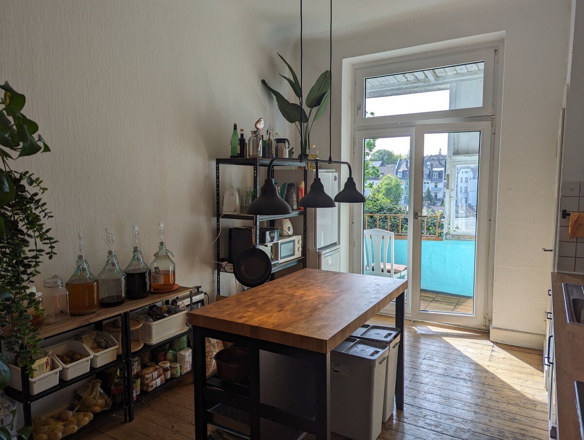 A light-filled kitchen features a wooden island workspace surrounded by open shelving containing various jars and pantry items. Two pendant lights hang above, while a large window and door lead to a balcony with green views, enhancing the bright atmosphere.