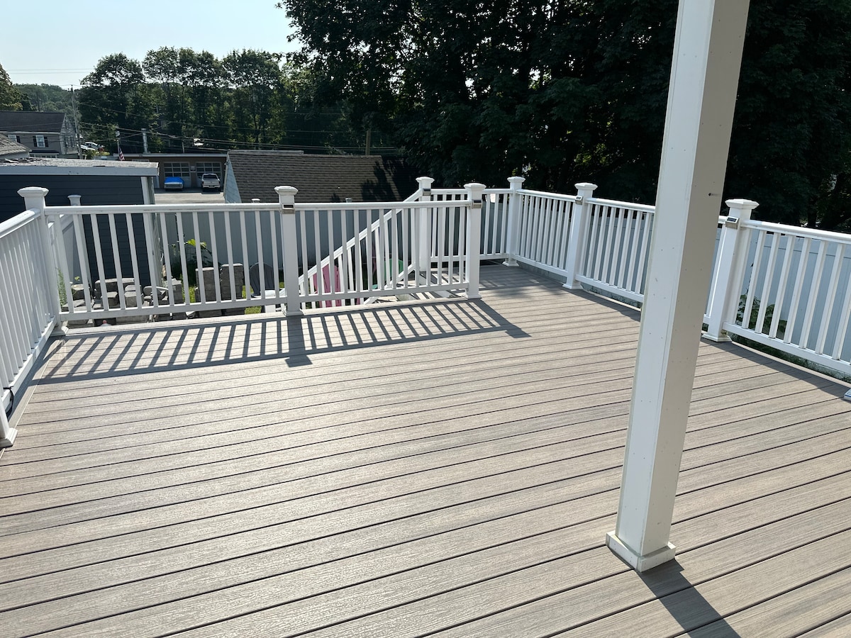 A spacious outdoor deck is presented with light-colored flooring and white railings. Sunlight casts shadows across the surface, highlighting the area for relaxation or gatherings. Trees and rooftops are visible in the background, offering a serene backdrop for outdoor activities.