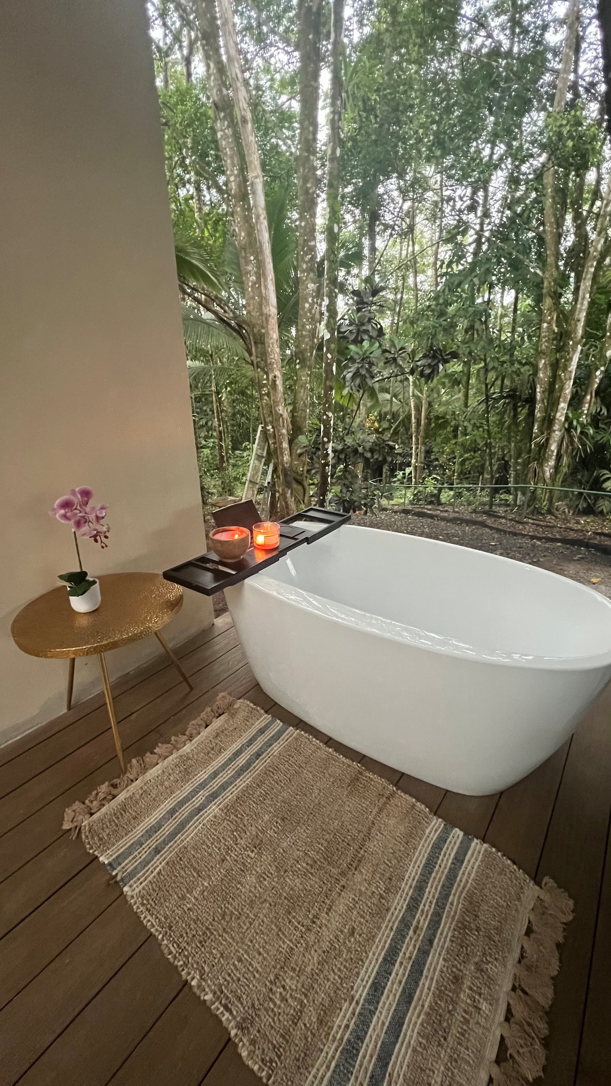 A modern freestanding bathtub is placed on a wooden deck, overlooking a lush jungle setting. A small, round side table holds lit candles and a purple orchid, enhancing the serene atmosphere. A woven rug lies beneath, adding textured warmth to the space.