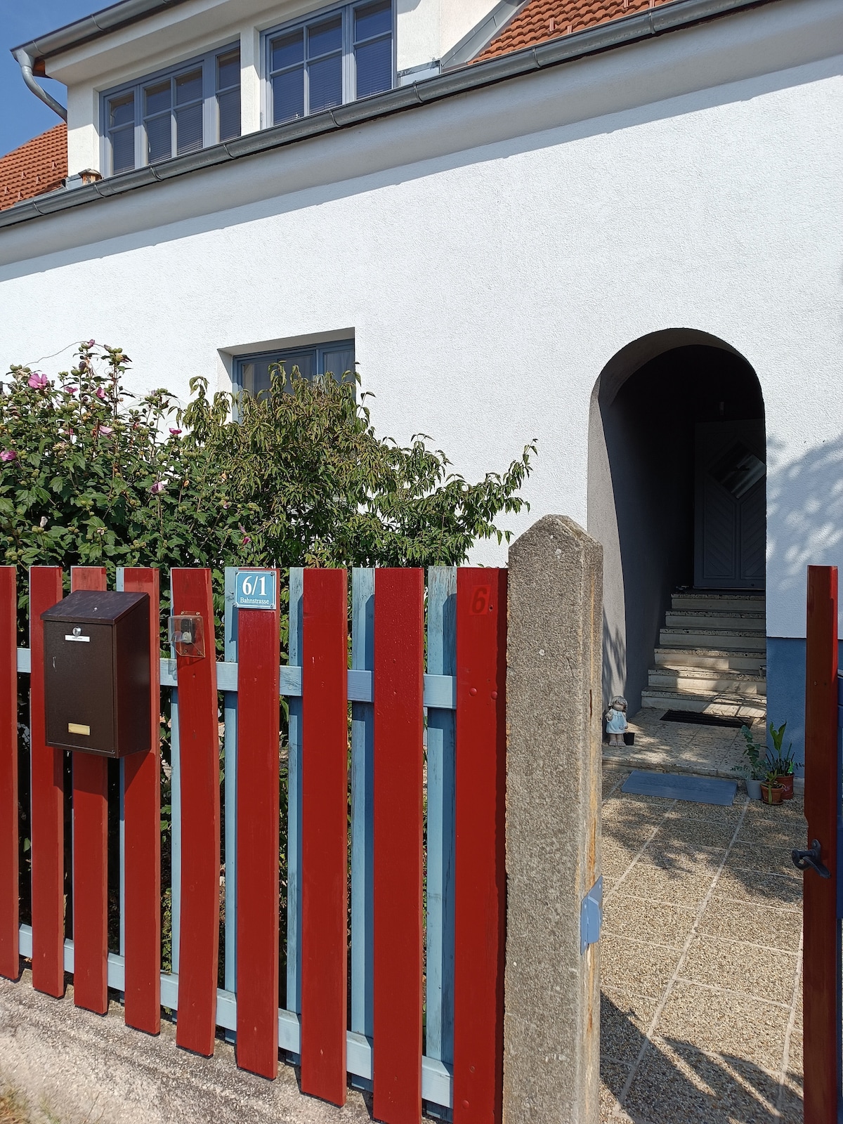A welcoming entrance is framed by a red and blue wooden fence, leading to a paved walkway. Lush greenery and flowering plants enhance the entrance, while a mailbox is positioned adjacent to the fence. The pathway guides guests toward the inviting arched doorway.