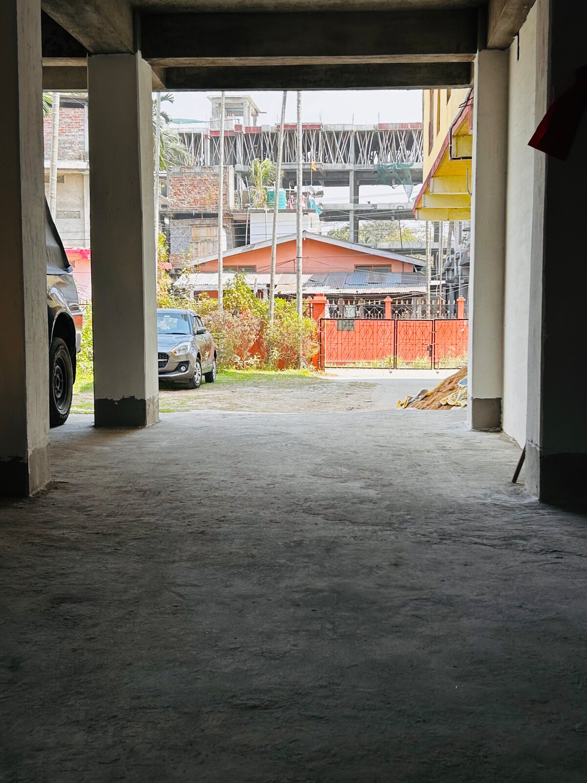 The image shows a view from an open area underneath a building, leading to an outdoor space. A pathway is visible along with parked vehicles, and colorful structures can be seen in the distance, providing a glimpse of the surrounding neighborhood.