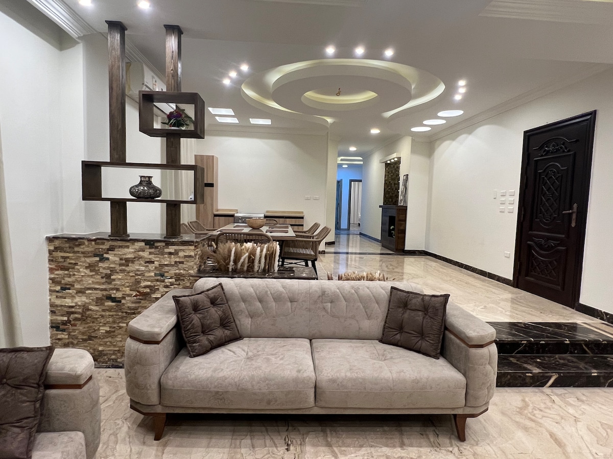 A spacious living area features a light-colored sofa with square cushions and a stone accent wall. Soft lighting highlights the coffered ceiling and the open layout leads to a dining space with wooden furniture, enhancing the room's ambiance.
