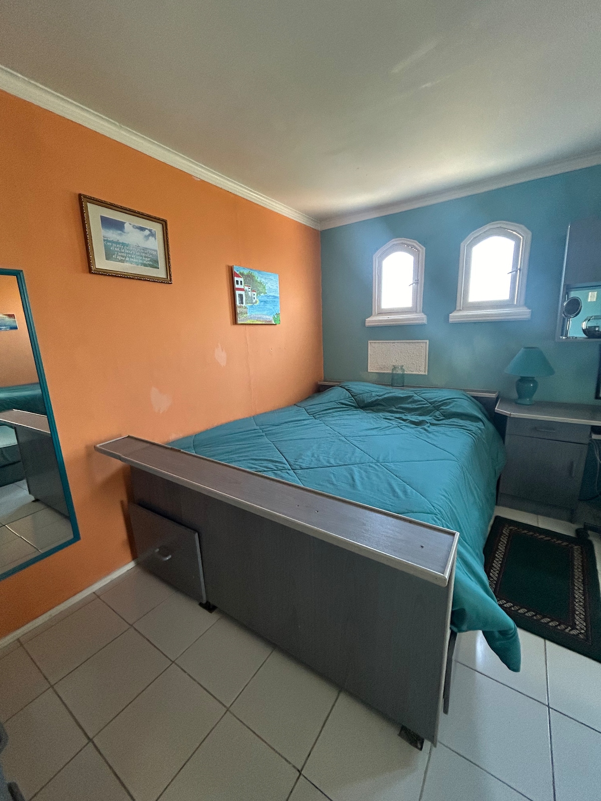 A bedroom features a spacious bed covered in a teal bedspread, complemented by orange and blue walls. Two small arched windows provide natural light. A mirror and a desk are visible, with a decorative painting enhancing the room's character.