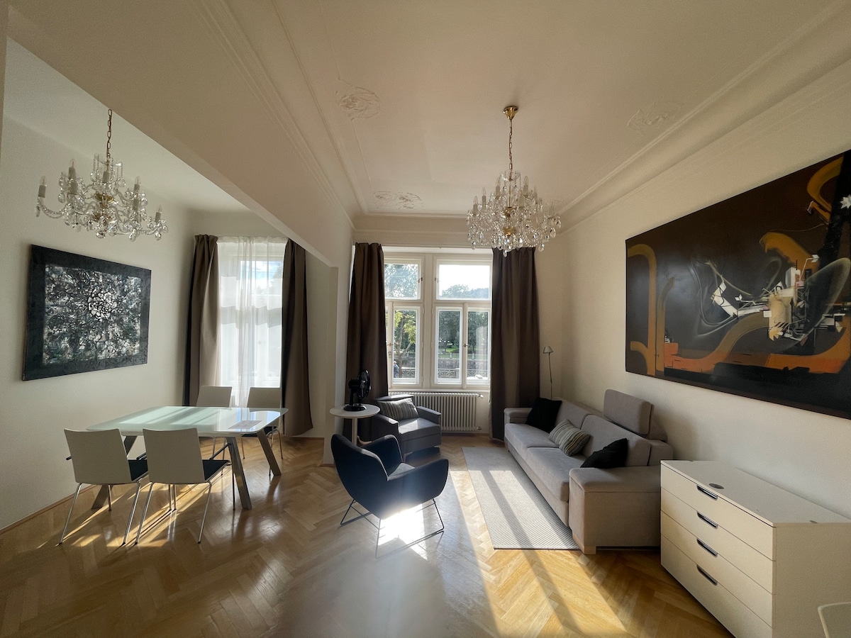 A spacious living area features a stylish combination of furniture, including a sectional sofa, armchair, and dining table with chairs. Elegant chandeliers hang from the ceiling. Large windows allow natural light to fill the room, enhancing the light-colored walls and hardwood floor.