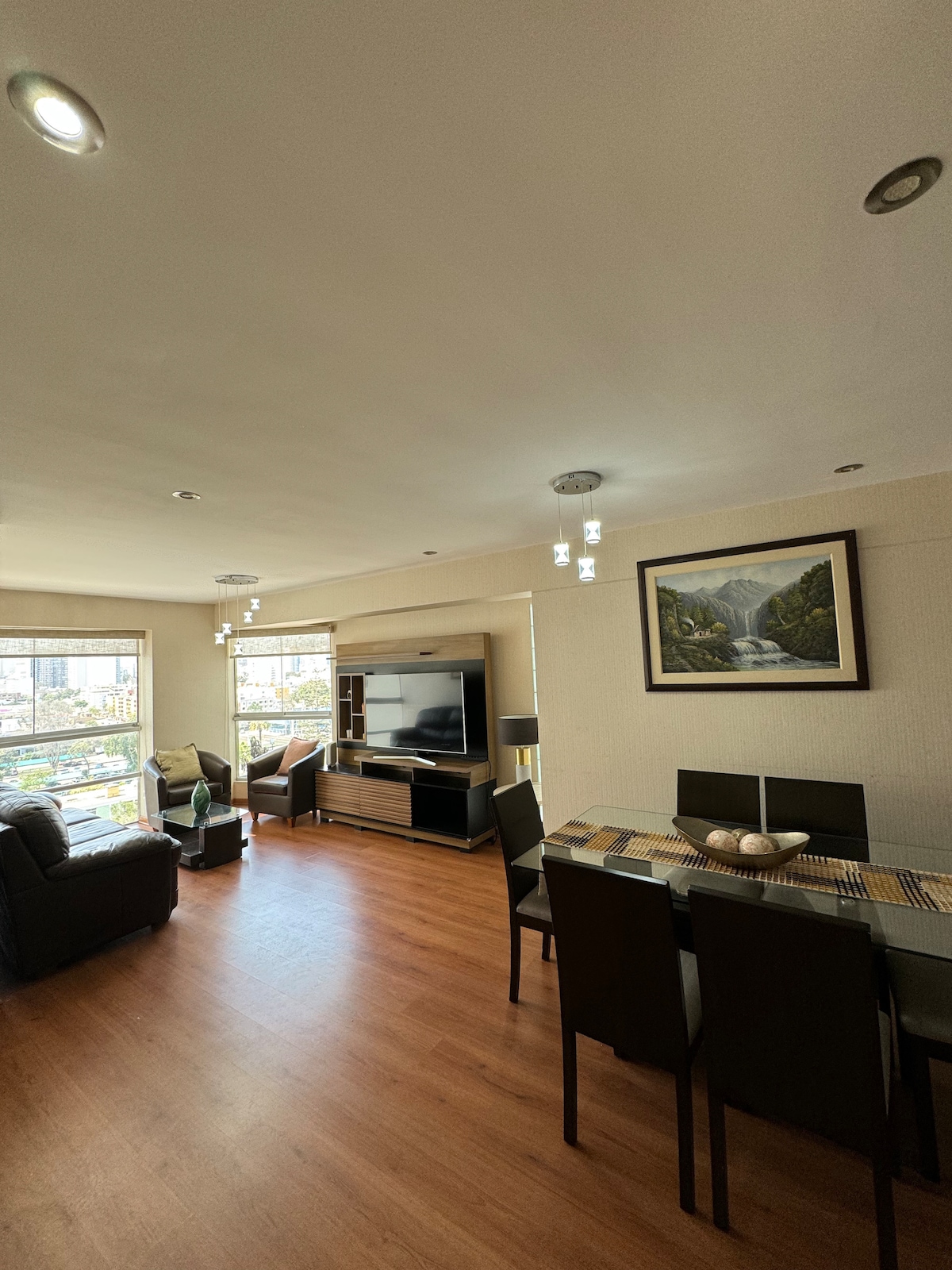 A spacious living area is visible, featuring a comfortable seating arrangement and a large television mounted on the wall. A dining table for six is positioned nearby, illuminated by natural light coming through two large windows with views of the outside.