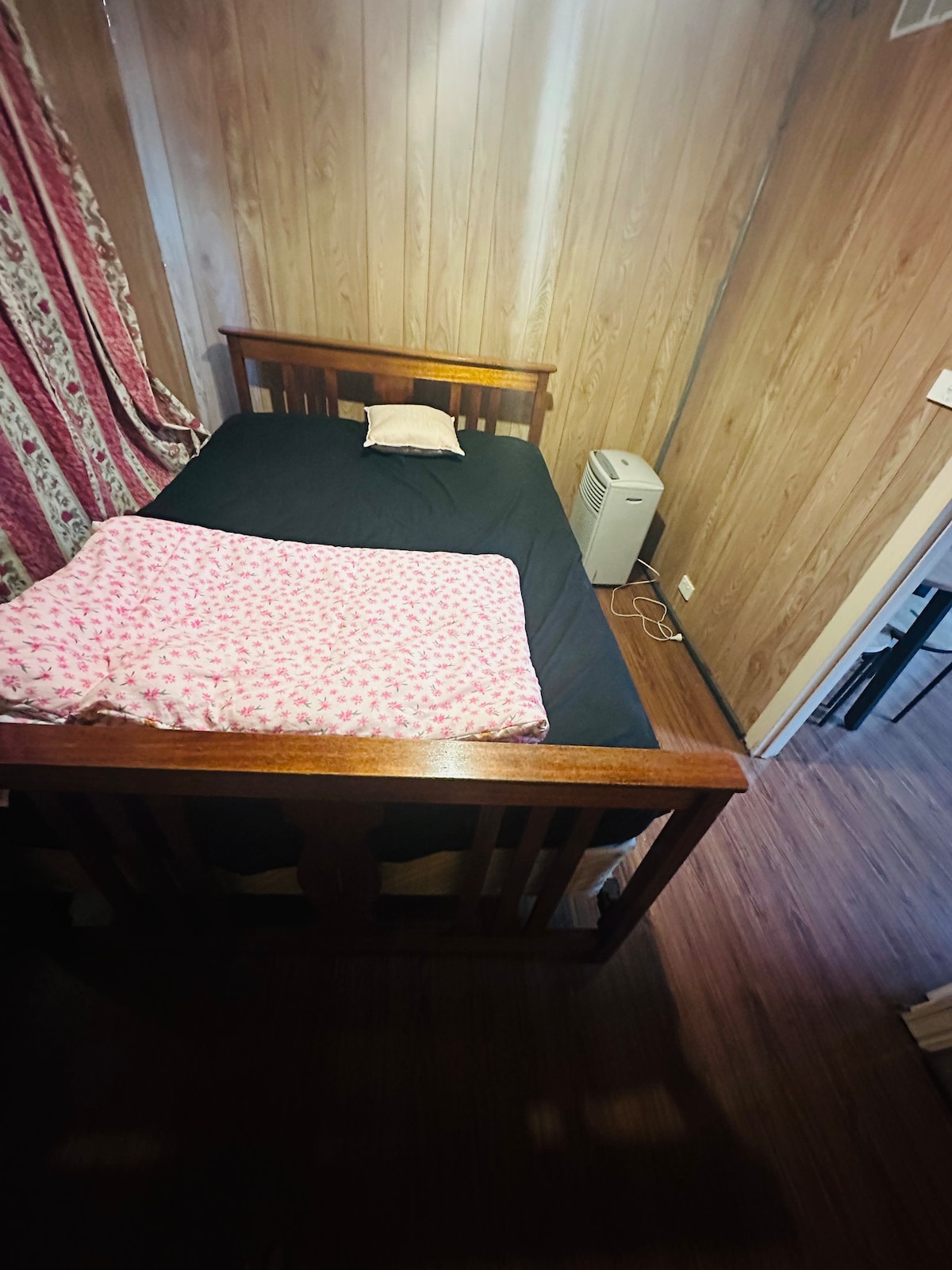 A wooden bed is positioned against the wall with a dark bedding set and a pink patterned blanket draped over it. A small pillow is placed at the head of the bed. A portable cooler is visible to the side, with wooden walls providing a natural aesthetic.