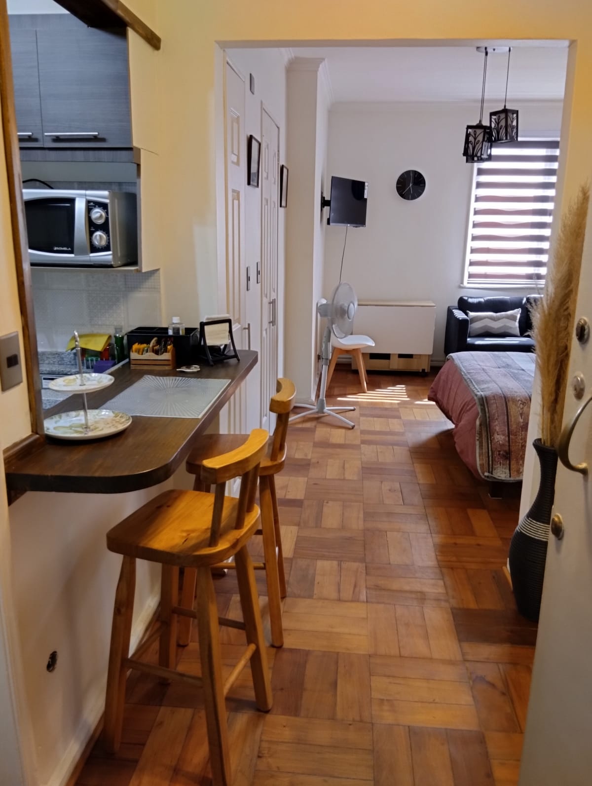 A view of the open layout showcasing a kitchenette with a microwave and a breakfast bar featuring two wooden stools. Natural light floods the space through a window, illuminating the cozy living area with a sofa and a television against the far wall.