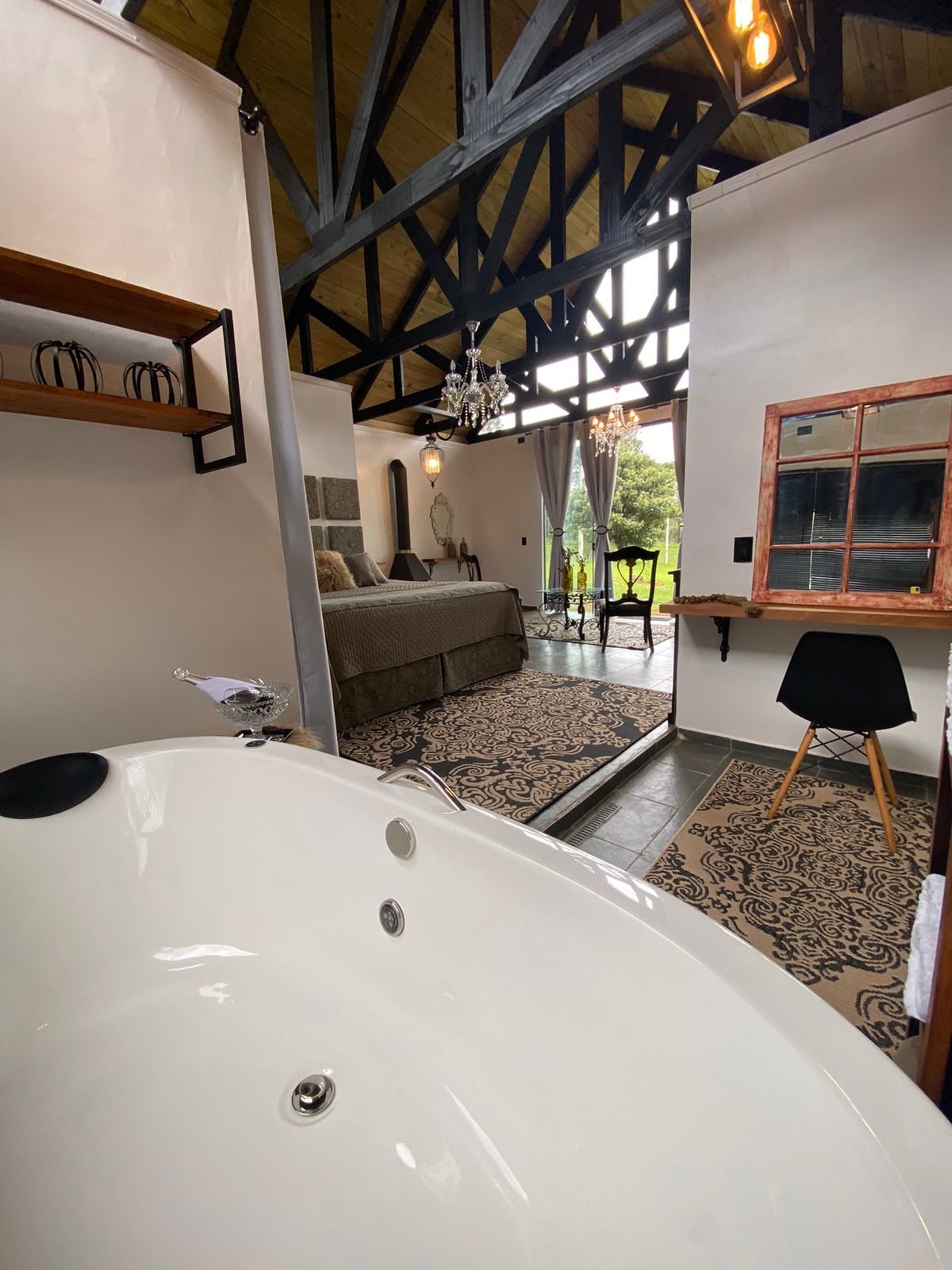 A spacious bathroom features a modern soaking tub and natural light filtering in from a nearby window. In the background, comfortable seating is visible along with a decorative rug. A contrast of rustic beams and contemporary furnishings creates a charming ambiance.