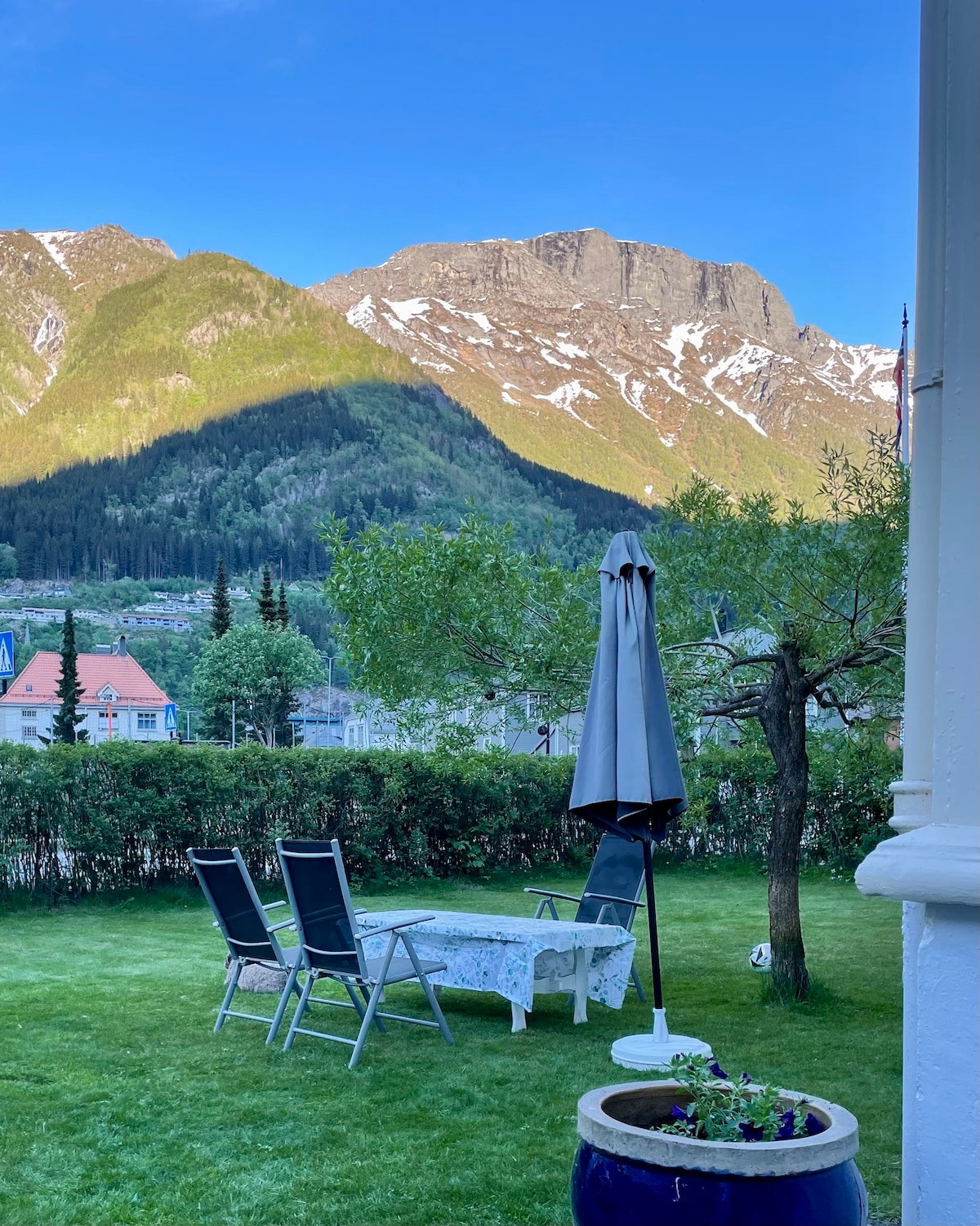 A serene outdoor area is featured, with a table surrounded by three chairs, all situated on well-kept grass. A large umbrella provides shade. In the background, mountains rise majestically, showing patches of snow and greenery under a clear blue sky.