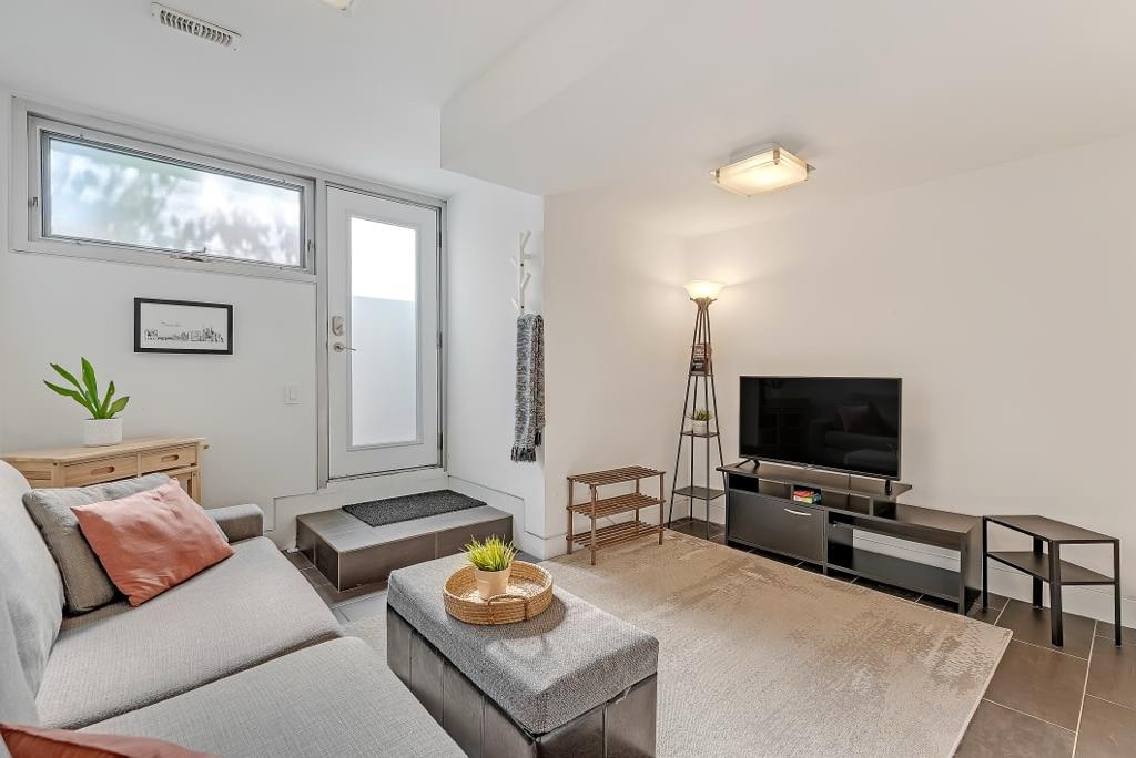 A comfortable living area is presented with a light-colored sectional sofa adorned with a decorative pillow. A small table holds a plant, while a sleek television stand sits opposite. Soft lighting is provided by a modern lamp, and large windows enhance natural light.