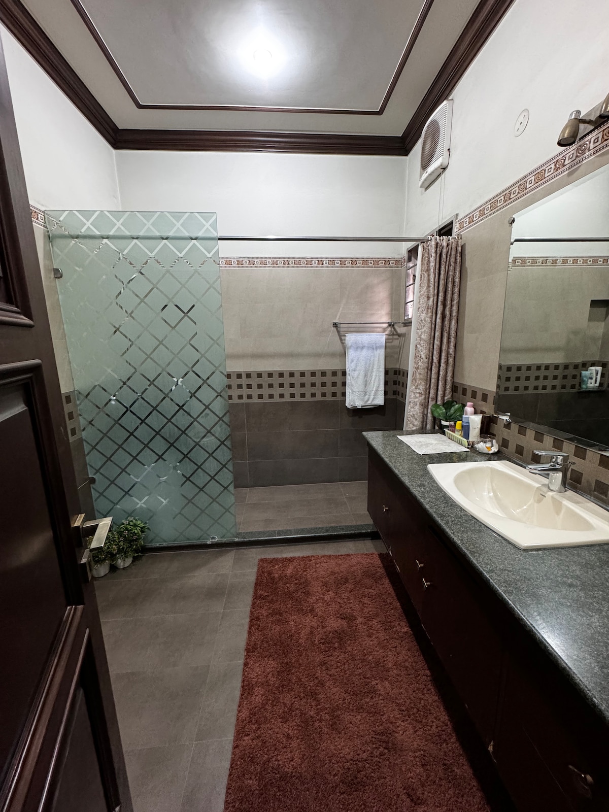 A bathroom features a glass shower enclosure with a textured design. A large mirror reflects the space above a dark countertop, accompanied by a small plant. A towel hangs from a bar next to a window, enhancing the room's functionality.