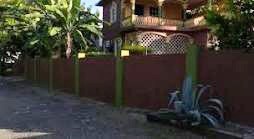 A charming two-story building is surrounded by a well-maintained fence, with tropical plants and trees enhancing the landscape. A clean pathway leads toward the entrance, highlighting the welcoming environment of the property.