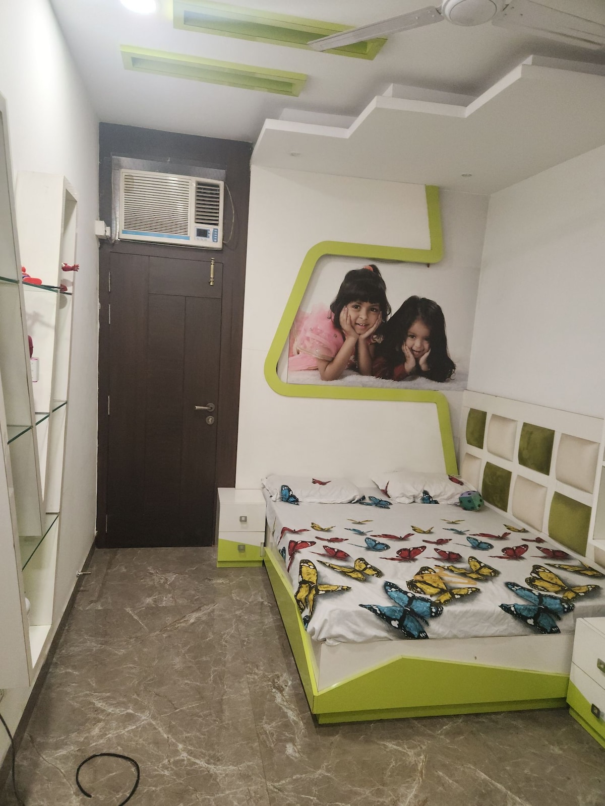A modern bedroom features a bed adorned with a butterfly-patterned blanket. The wall behind the bed is decorated with a colorful mural, while a sleek shelf provides space for personal items. An air conditioning unit is mounted above the door, facilitating comfort.
