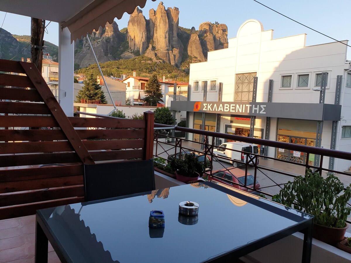 A patio table with a glass top is positioned next to a wooden swing chair. Lush greenery is placed around the space, while a striking rock formation is visible in the background, complemented by buildings along the street.