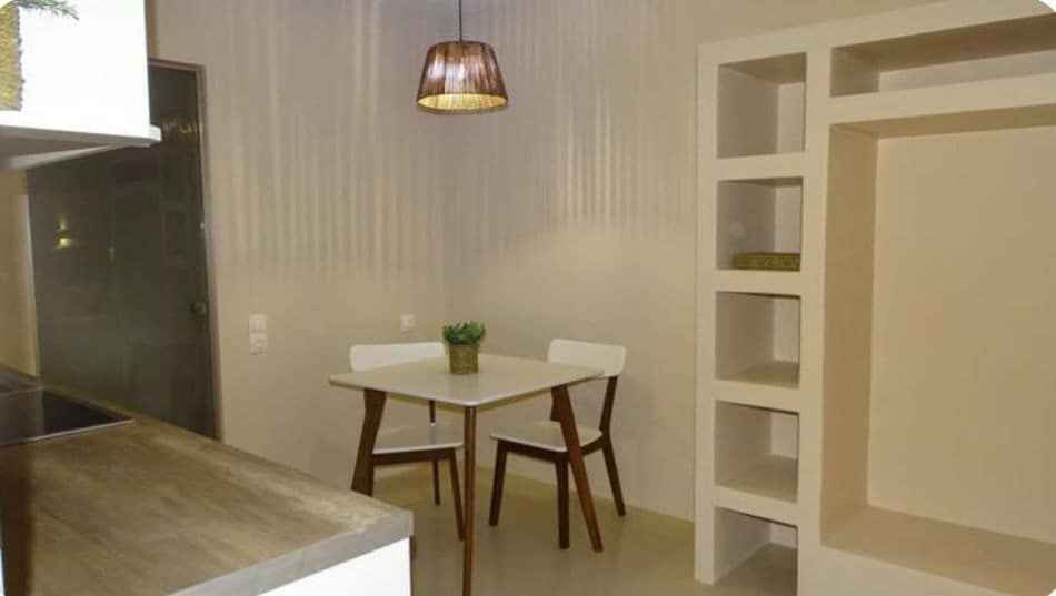 A simple dining area features a white table surrounded by two wooden chairs. Soft light is provided by a pendant lamp hanging above the table. A small potted plant adds a touch of greenery, while a shelving unit is visible nearby.