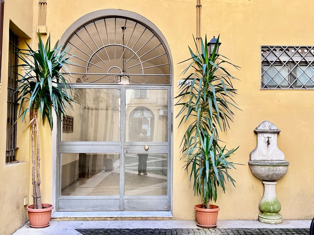A large arched entrance door is framed by two potted plants on either side. The door features a round window design above it, leading into a well-lit interior space with a stone fountain visible nearby, all set against a warm yellow wall.