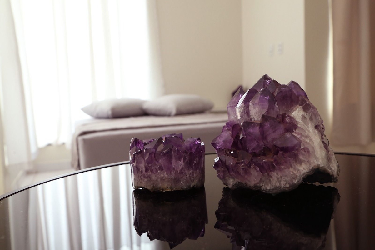 A sleek black table features two clusters of amethyst crystals, showcasing their rich purple hues. In the background, a spacious bed with soft grey pillows is slightly visible, framed by light, sheer curtains that allow natural light to brighten the area.