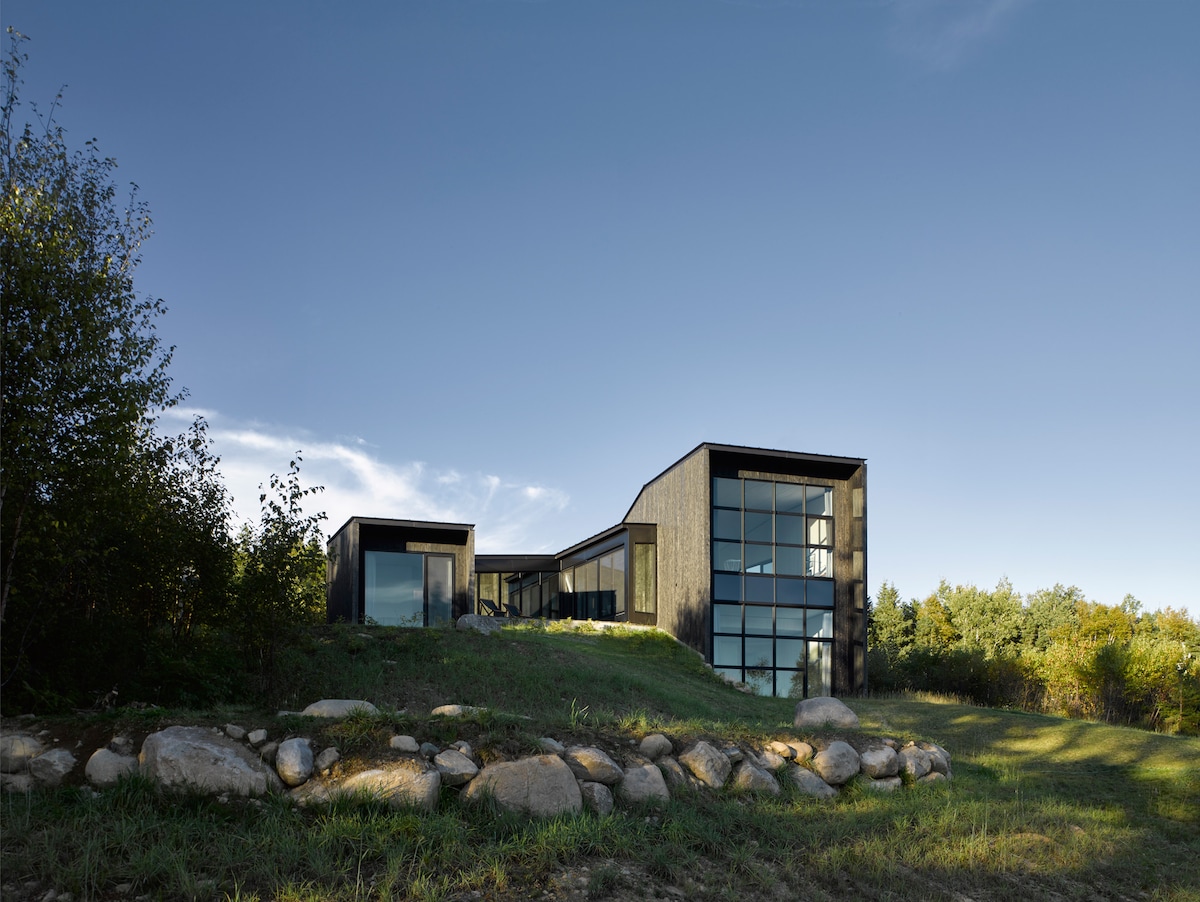A modern structure with a unique design is set against a clear blue sky. Large windows reflect the surrounding greenery, while a natural stone border defines the property’s edge. Lush landscaping adds to the serene outdoor environment.