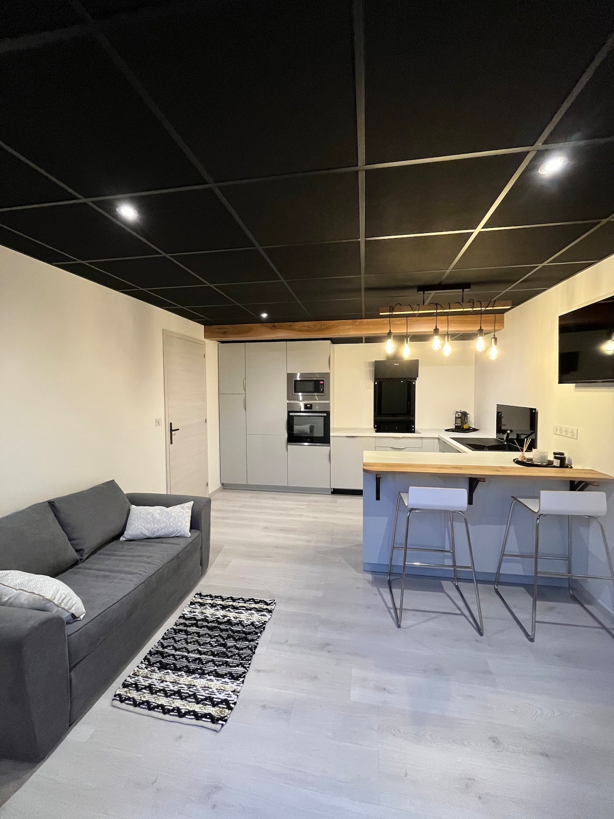A modern living area is observed, featuring a comfortable gray sofa and a patterned rug. A compact kitchen with sleek cabinetry and appliances is visible, complemented by a small dining area with two stools. The ceiling showcases stylish pendant lighting and the floor is finished in light wood.