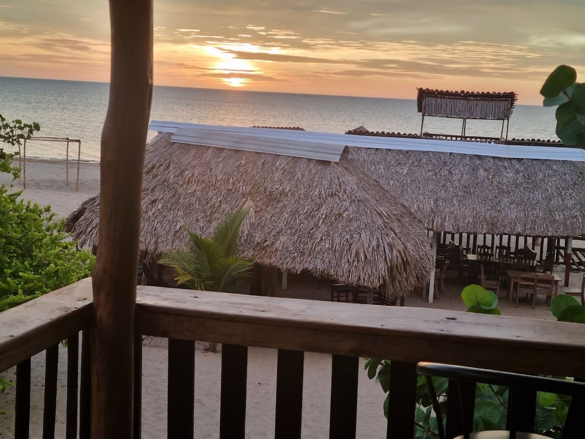A serene beach view is captured at sunset, showcasing the horizon over calm waters. Thatched-roof structures are visible in the foreground, surrounded by sandy beach and lush greenery, creating a natural and tranquil setting.