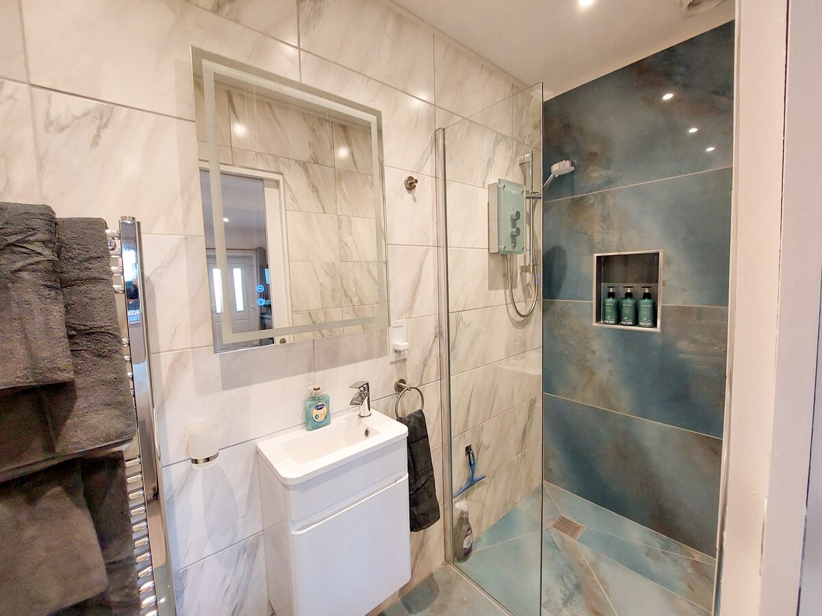 A modern shower room features tiled walls in neutral tones, a shallow sink with a mirror above, and a glass-enclosed shower with a rainfall showerhead. A stylish towel rack is visible, alongside a shelf displaying decorative bottles.