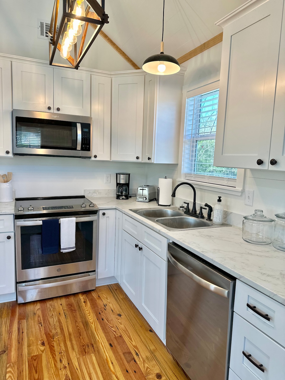A modern kitchen is presented with white cabinetry and marble countertops. Stainless steel appliances, including a microwave and a dishwasher, are neatly arranged. A coffee maker, sink, and various kitchen essentials enhance the functionality. Natural light streams through a window, illuminating the warm wood flooring.