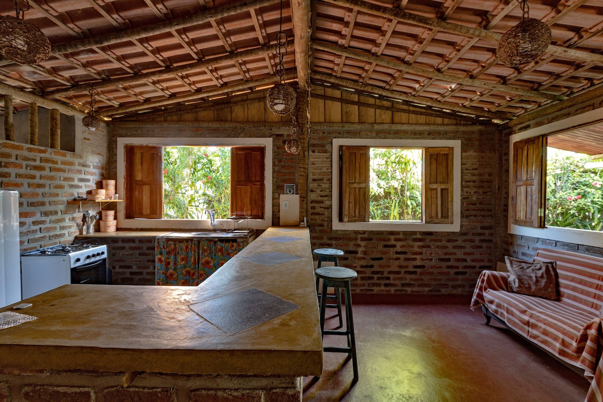 An inviting kitchen area is showcased, featuring a rustic design with exposed brick walls and a vaulted ceiling. Large windows allow natural light to fill the space, revealing views of the greenery outside. A kitchen counter with bar stools and cooking appliances is visible, complemented by a cozy seating area.