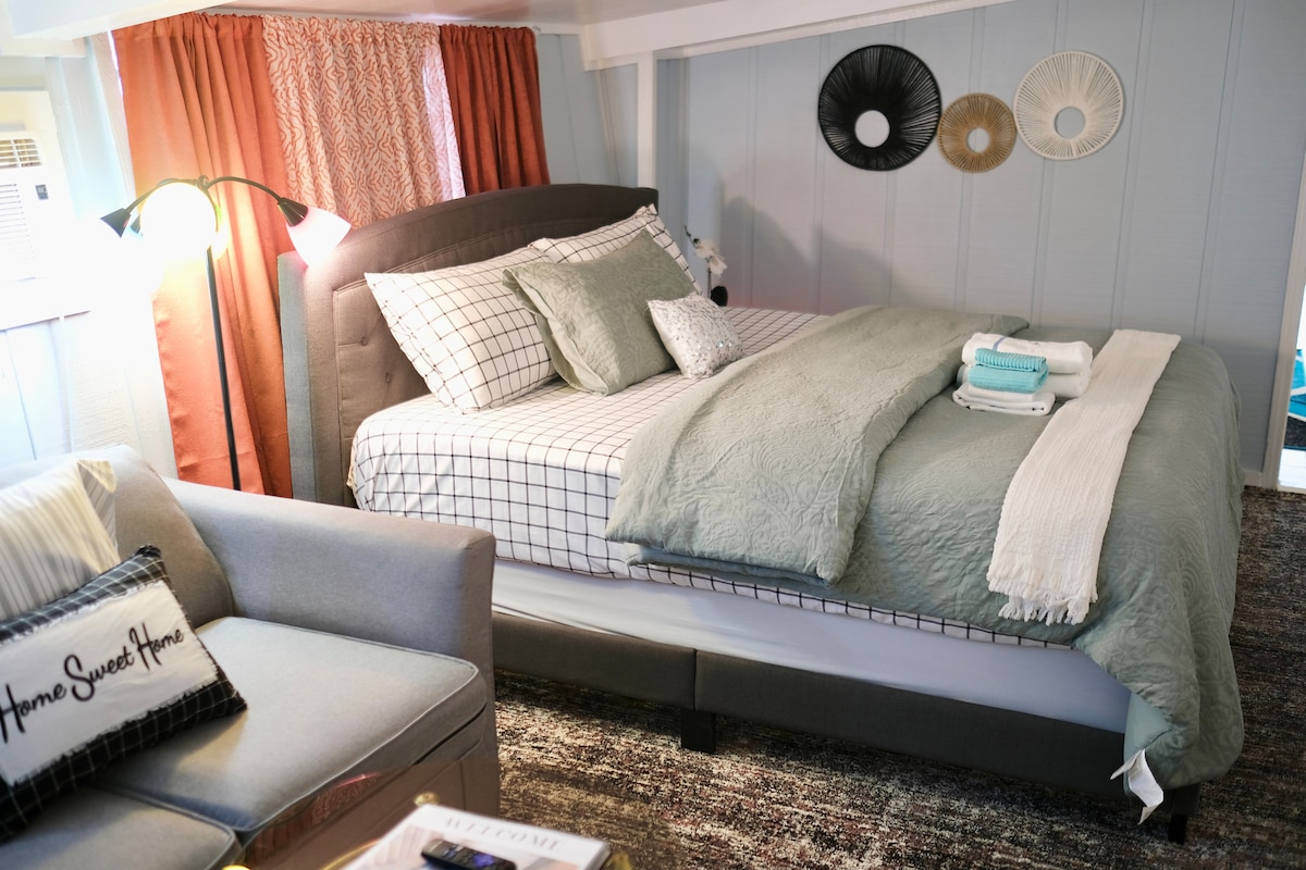 A cozy bedroom features a spacious bed with a soft, textured blanket layered on top. Stylish pillows adorn the bed, while a comfortable chair sits nearby. Decorative wall accents in neutral tones add warmth, complemented by natural light filtering through the window with soft curtains.