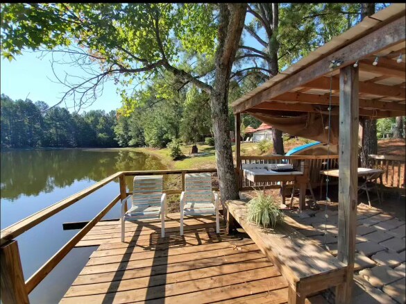 A wooden deck overlooks a serene pond, surrounded by lush trees. Two chairs are positioned beside a table, offering a space for relaxation. The area is enhanced by warm sunlight and natural scenery, creating a restful outdoor setting.