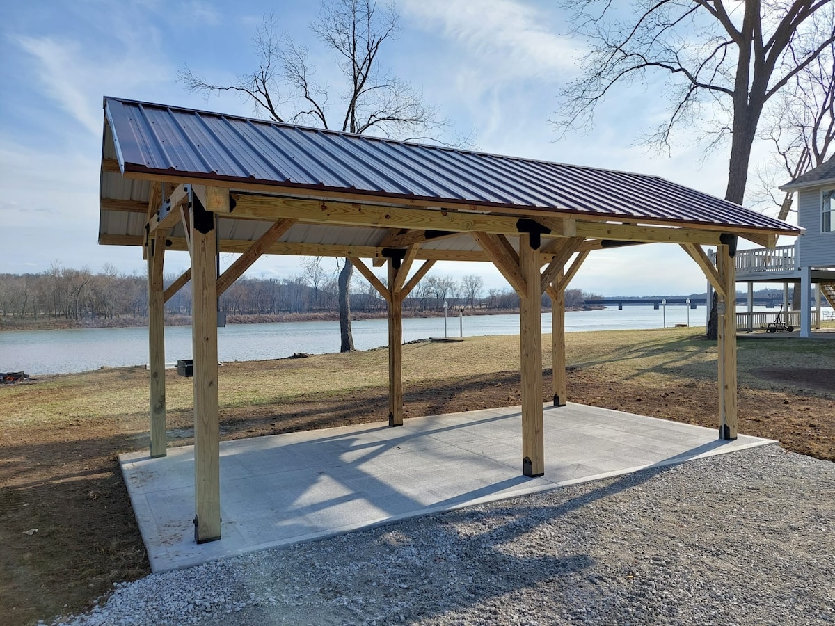 A covered shelter is positioned on a concrete pad, featuring wooden beams supporting a metal roof. The structure overlooks the riverside, with a view of the water and distant trees, providing a shaded area ideal for outdoor gatherings.