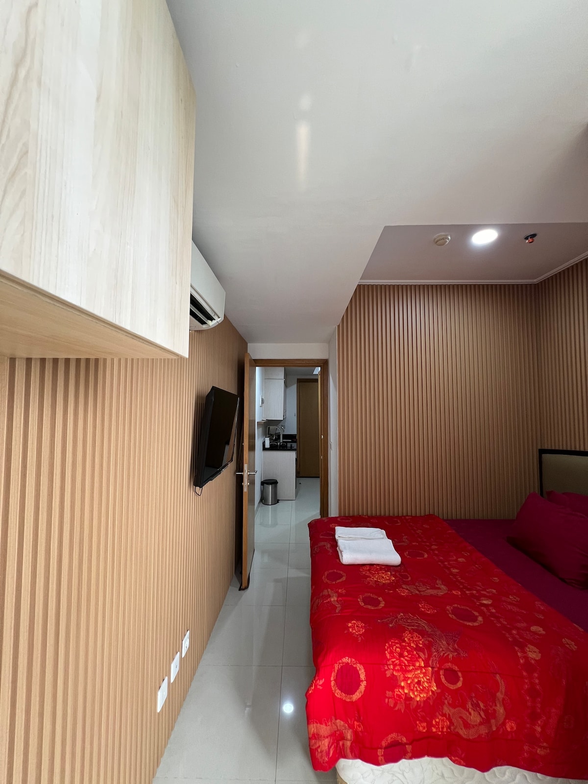 A cozy bedroom is highlighted by textured wood paneling and soft lighting. A spacious bed with a bright red coverlet is positioned against the wall, complementing a flat-screen TV mounted nearby. A doorway leads to a modern kitchen area visible in the background.