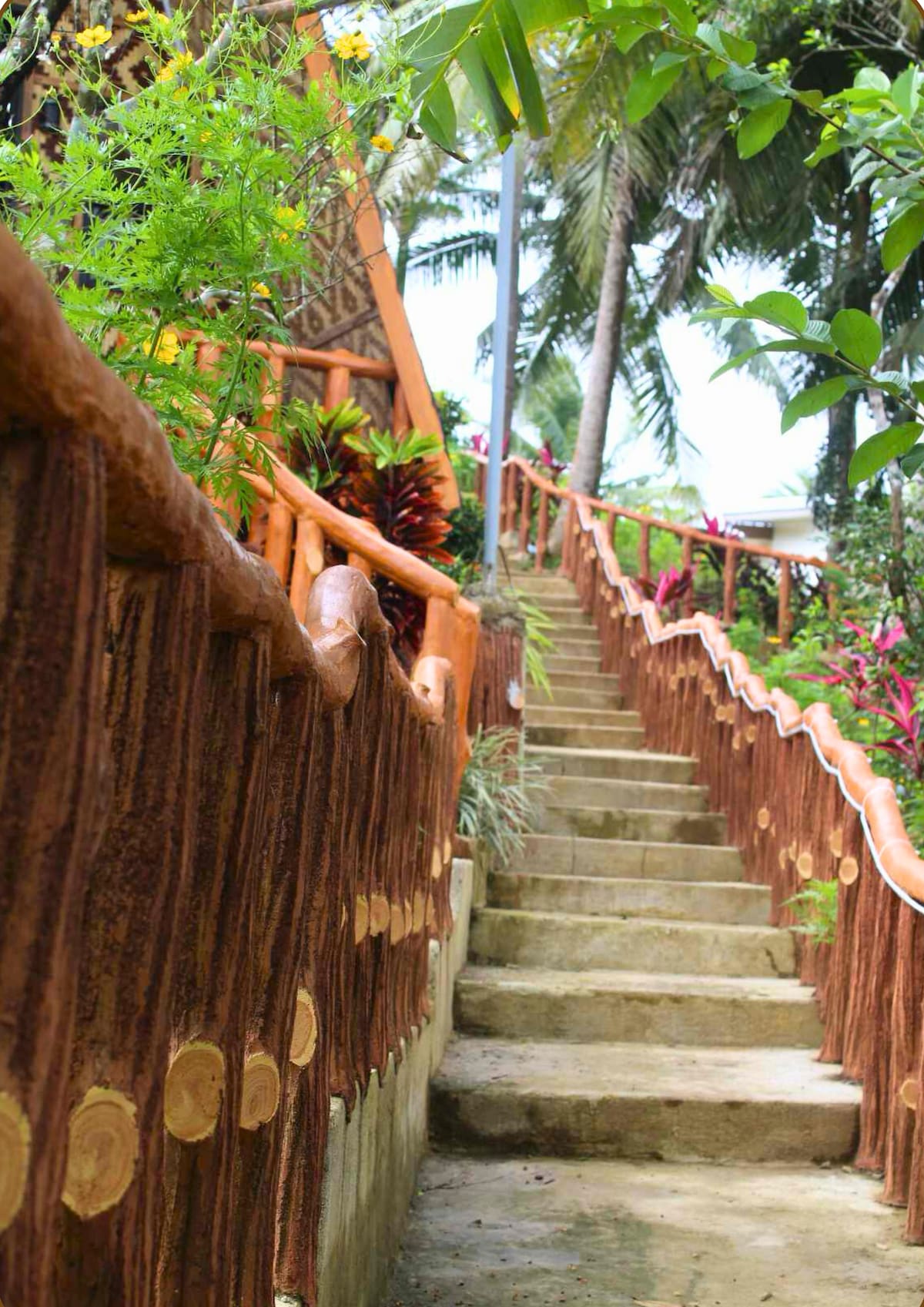 A winding stone staircase is framed by a textured wooden railing and bordered by lush greenery. Vibrant tropical plants are visible along the sides, leading up through a peaceful garden area towards the house.