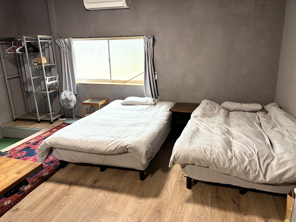 The room contains two spacious beds, each dressed in light-colored bedding. A window with light curtains lets in natural light, and a fan is positioned nearby. A wooden nightstand and a small shelf stand against the wall, providing additional space for personal items.