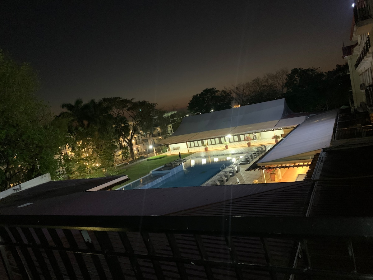 A serene night view of a swimming pool illuminated softly. Surrounding the pool, lounge chairs are arranged for relaxation. The hotel area features trees and well-maintained greenery, contributing to a calm atmosphere under the darkening sky.