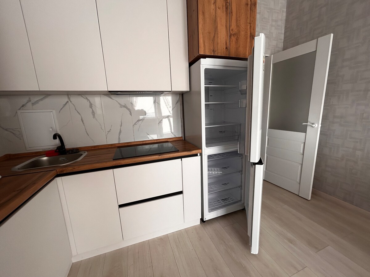 The kitchen features sleek white cabinetry complemented by wooden accents. A modern countertop includes a built-in stovetop beside a small sink. The open refrigerator stands with its door ajar, revealing ample storage space, while a neutral-tone wall creates a bright atmosphere.